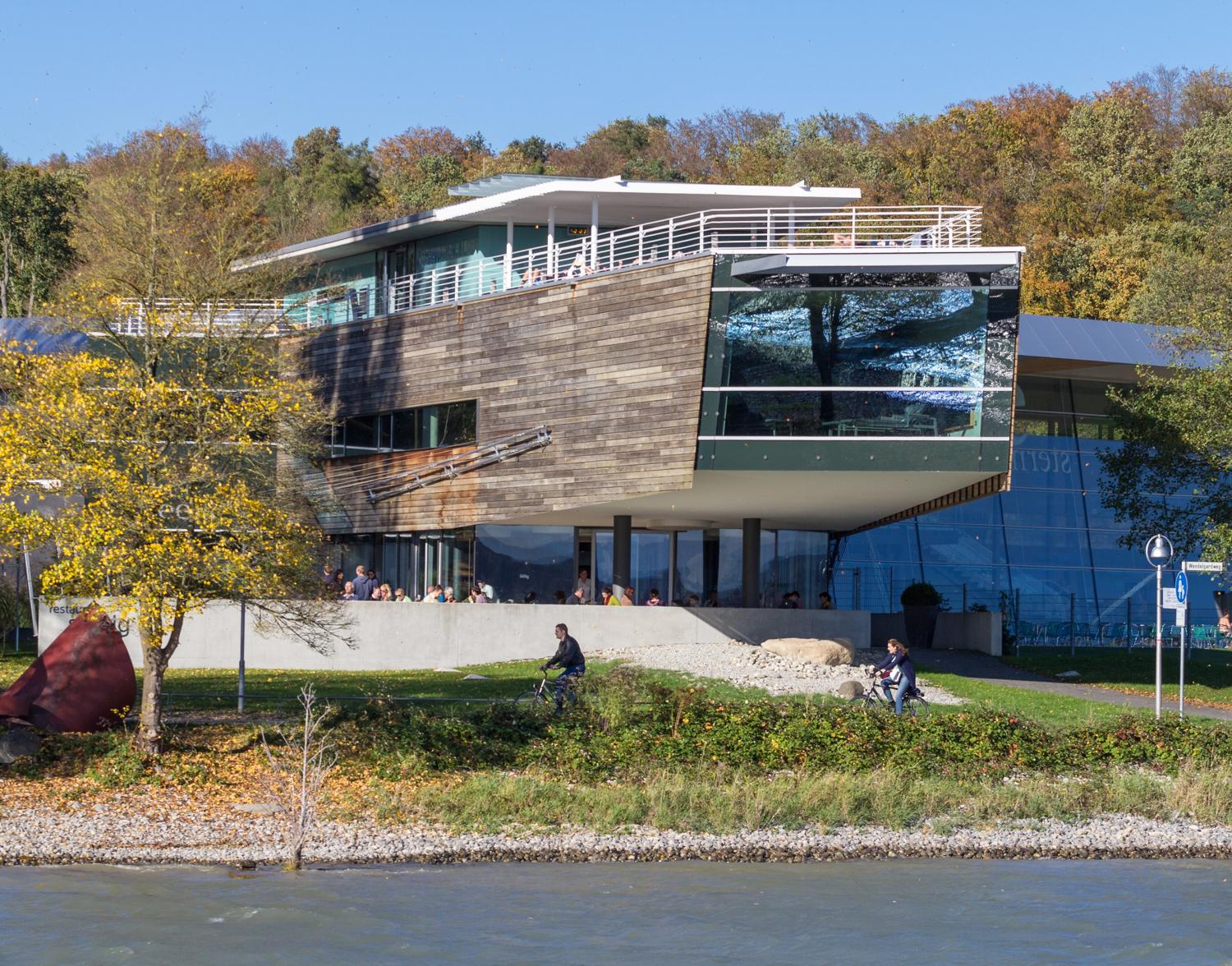 Die Bodensee-Therme Konstanz vom Wasser aus: Bau aus Holz und Glas mit Terrasse, davor Uferweg, Herbstbäume und der See