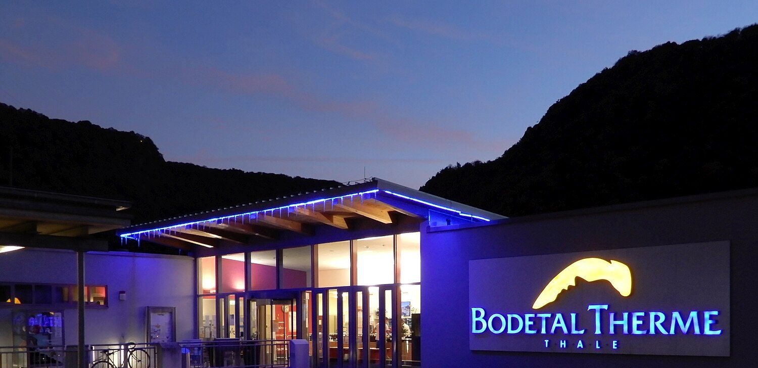 Eingang der Bodetal Therme in Thale in der Dämmerung: blau beleuchtetes Vordach, leuchtender Schriftzug, dahinter die dunklen Harzberge