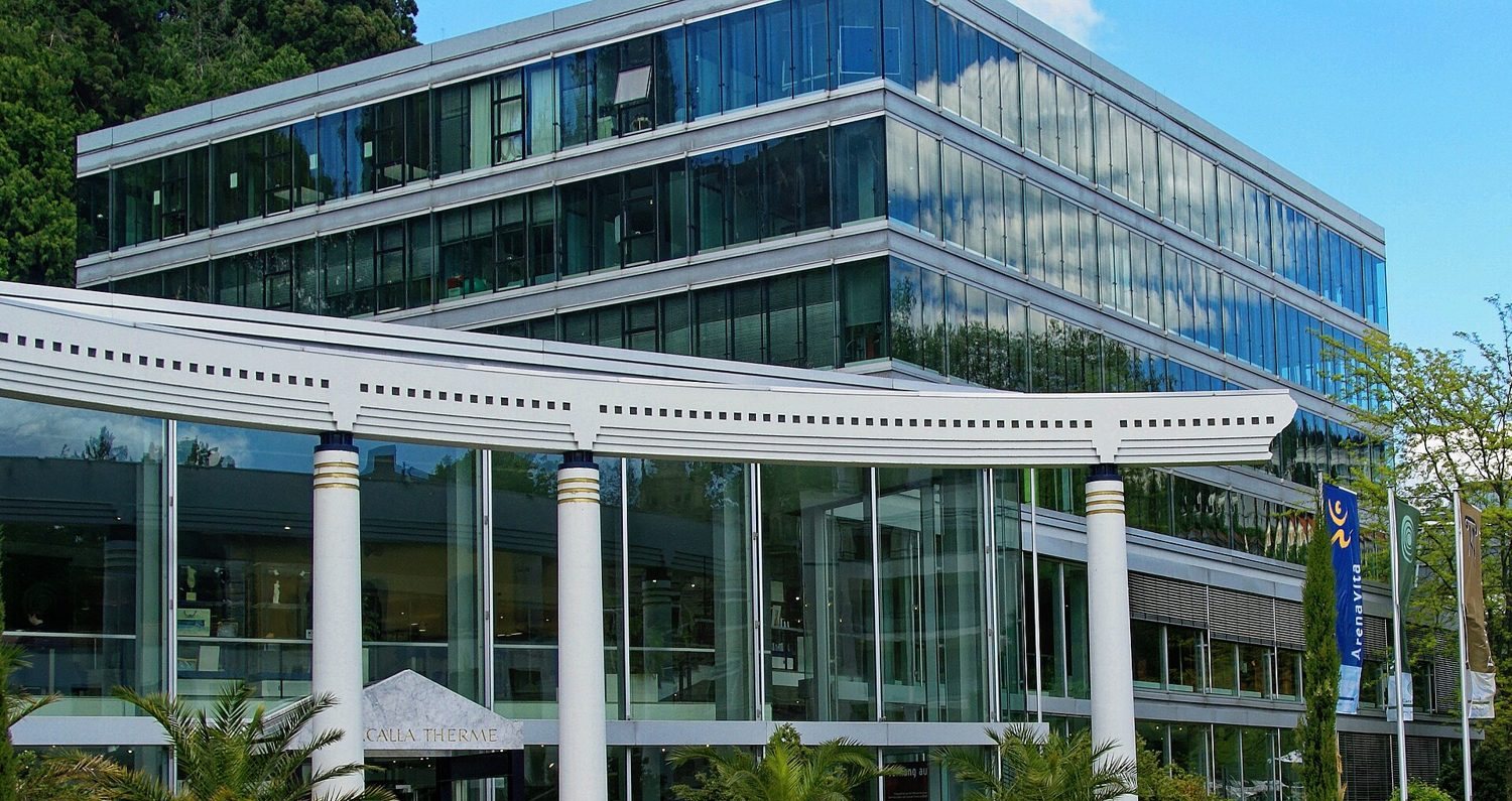 Eingang der Caracalla Therme in Baden-Baden: geschwungenes weißes Vordach auf Säulen vor einer Glasfassade, darüber blauer Himmel
