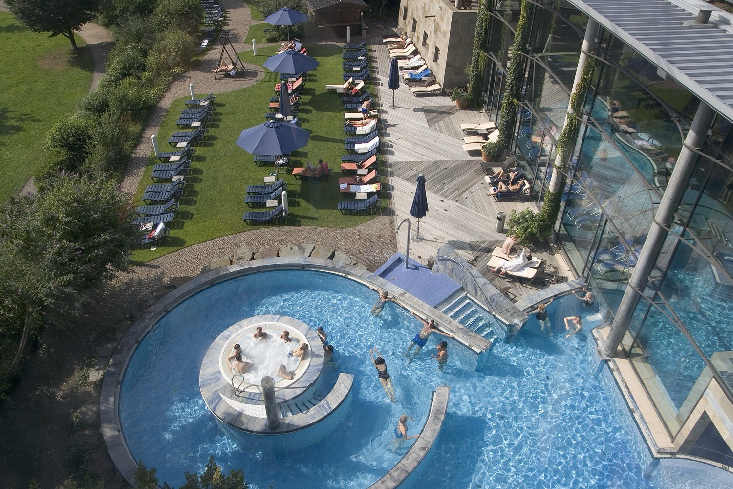 Außenbecken der Claudius Therme in Köln von oben: rundes Thermalbecken mit Whirlpool-Insel, Badegäste im türkisblauen Wasser, daneben Liegewiese mit Sonnenschirmen