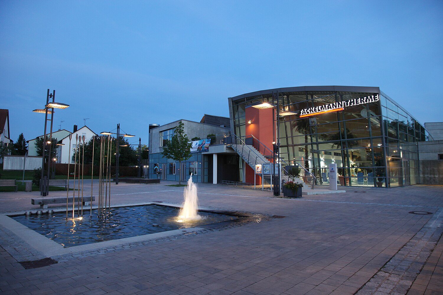 Die Fackelmann Therme in Hersbruck in der Abenddämmerung: beleuchtete Glasfassade, davor ein Brunnen mit Wasserfontäne