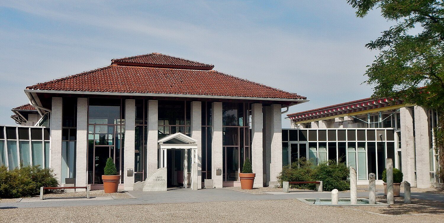 Eingangsgebäude der Limes-Thermen in Aalen: Pavillon mit Ziegeldach und Säulen, davor ein Kiesplatz mit Pflanzkübeln