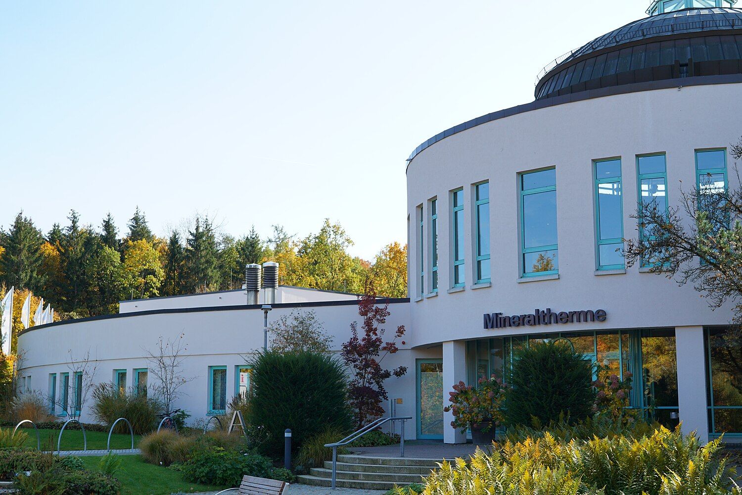 Eingang der Mineraltherme Böblingen: heller Rundbau mit türkisfarbenen Fenstern und Kuppel, davor Farne und herbstlich gefärbter Wald