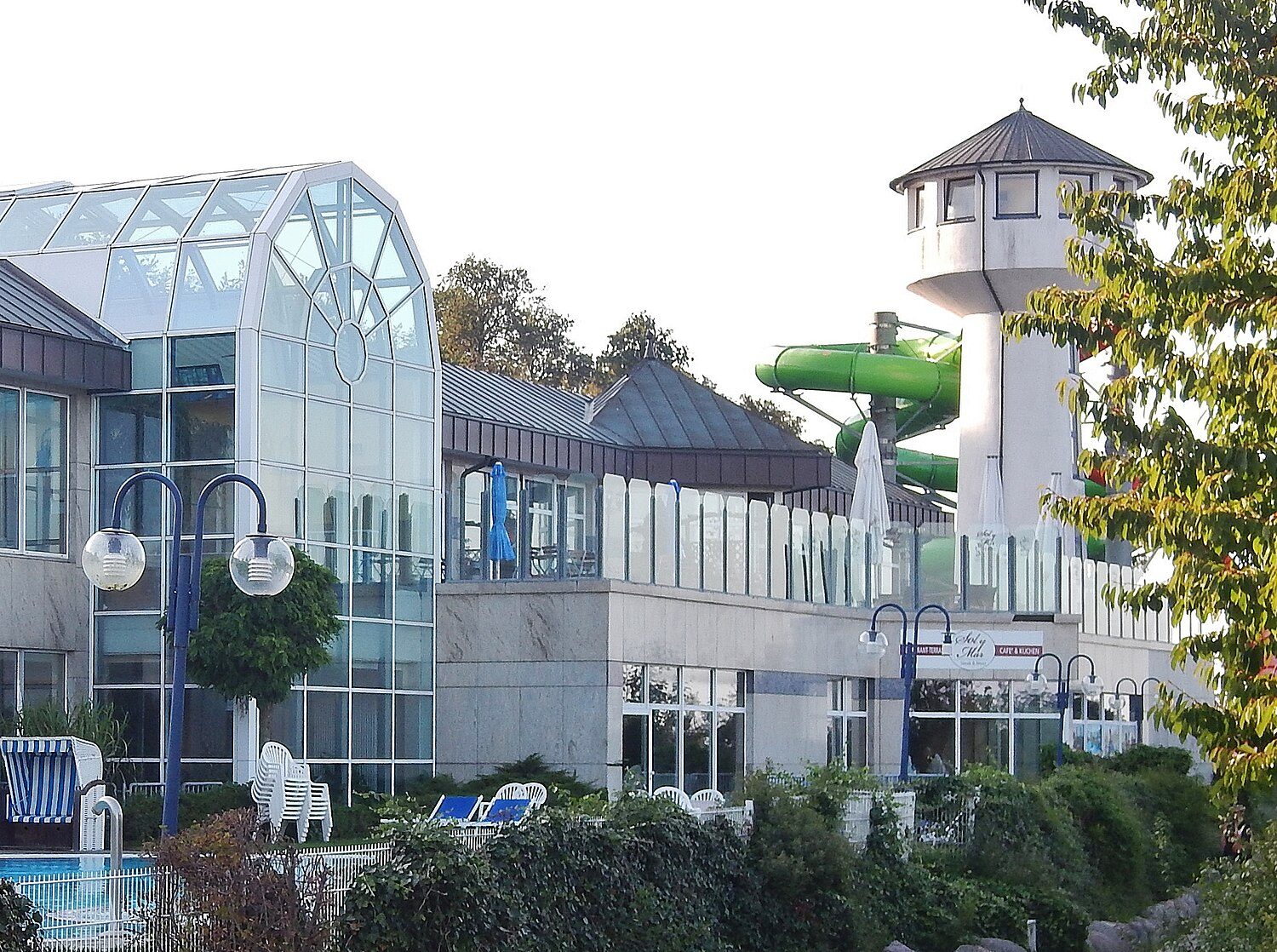 Die Ostsee Therme in Scharbeutz von außen: weiße Glasgiebel, ein Turm mit grüner Röhrenrutsche, davor Strandkörbe und Hecken