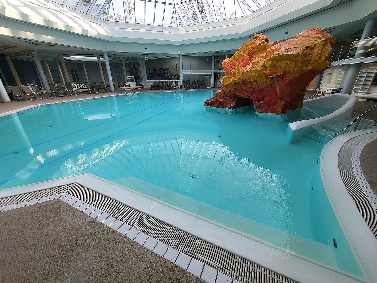 Innenbecken der OstseeTherme Usedom: türkisfarbenes Wasser unter einer Glaskuppel, ein orange-roter Felsen mit kleiner Rutsche, Liegen am Beckenrand