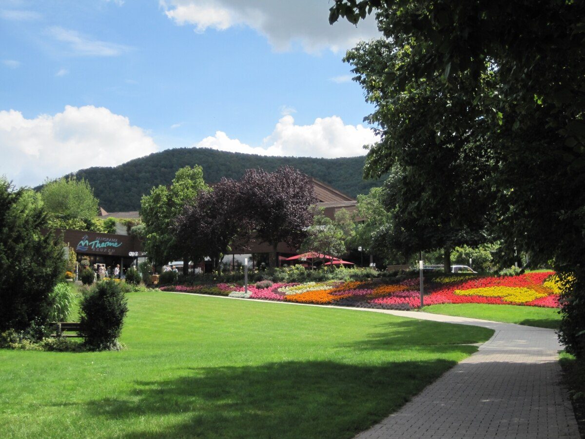 Kurpark vor der Panorama Therme Beuren: Blumenbeete und Rasen, dahinter der Eingang der Therme und ein bewaldeter Höhenzug