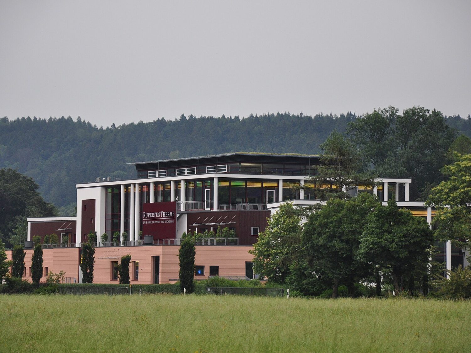 Die RupertusTherme Bad Reichenhall mit weißen Säulen, Dachterrasse und Schriftzug, davor eine Wiese, dahinter der Wald