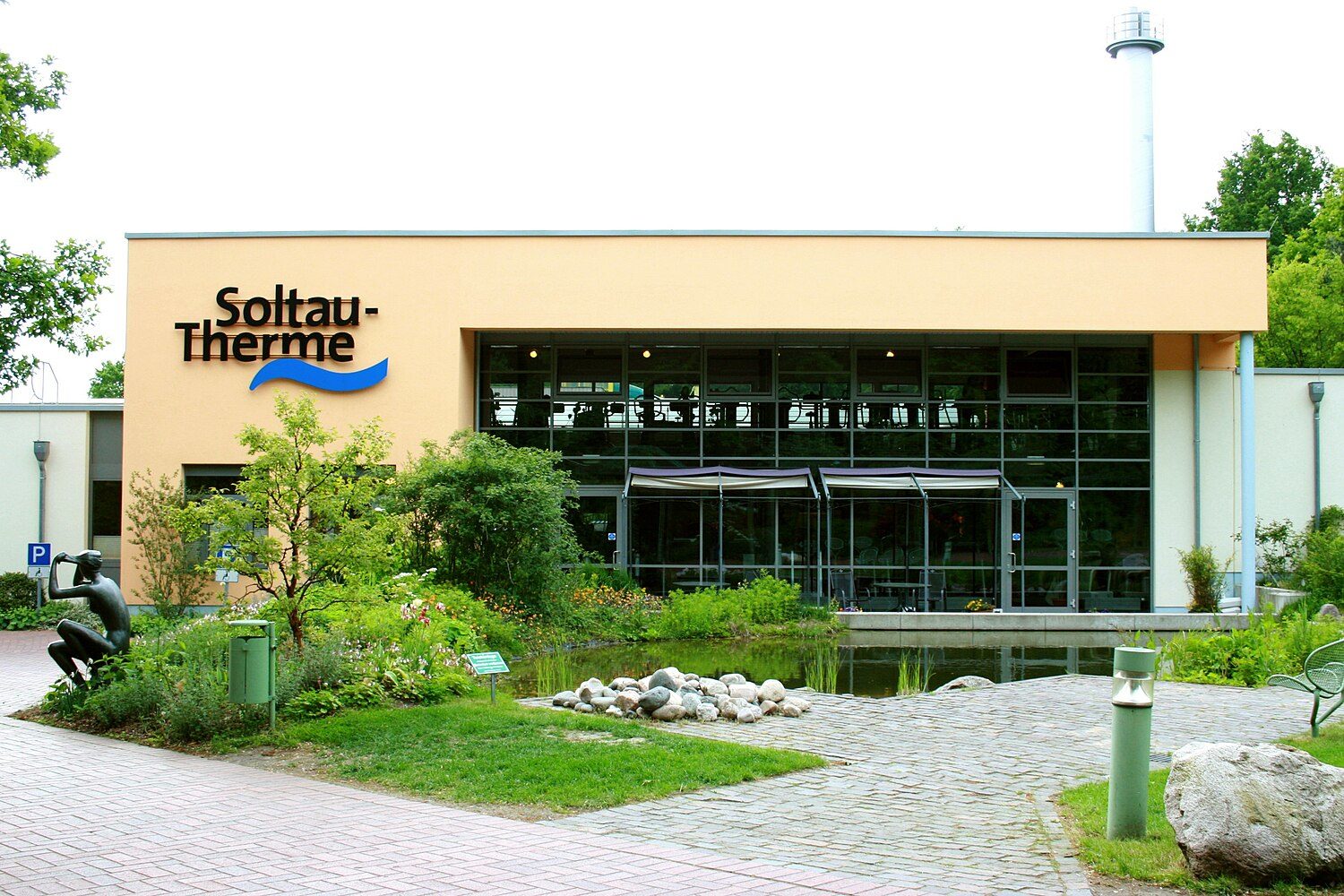 Eingangsbereich der Soltau Therme: heller Bau mit Glasfront und Schriftzug, davor Teich, Findlinge und Grün