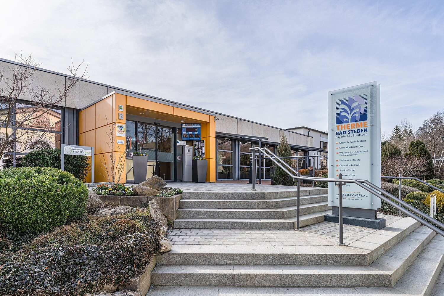 Eingang der Therme Bad Steben: flacher Bau mit orangefarbenem Portal, Stufen und Bepflanzung, davor die Stele mit dem Logo