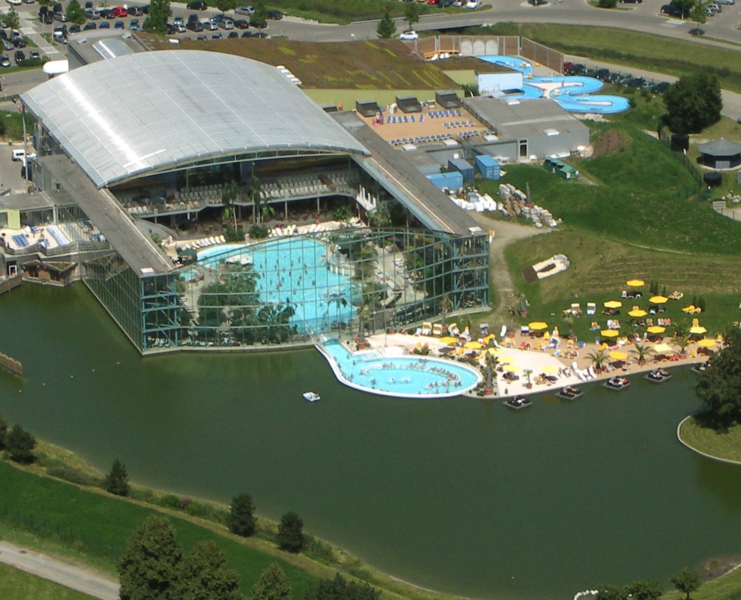 Luftaufnahme der Therme Bad Wörishofen: gläserne Palmenhalle mit türkisfarbenem Becken, davor das runde Außenbecken, Liegewiese mit gelben Sonnenschirmen und ein See