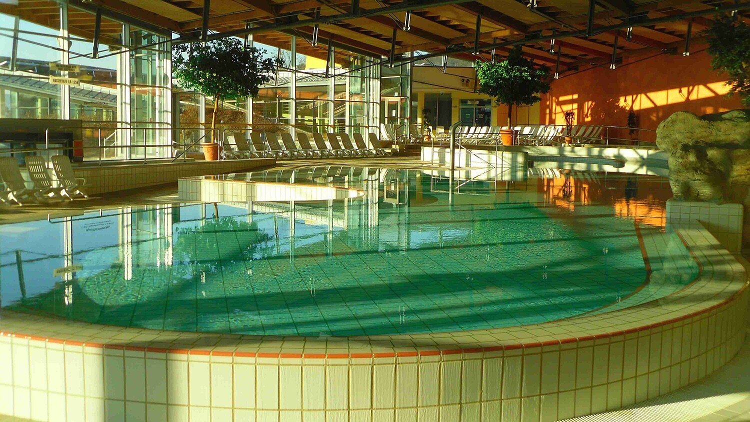 Badehalle der Therme Obernsees im Abendlicht: geschwungenes Becken mit türkisfarbenem Wasser, Liegen an der Glasfront, Holzdecke