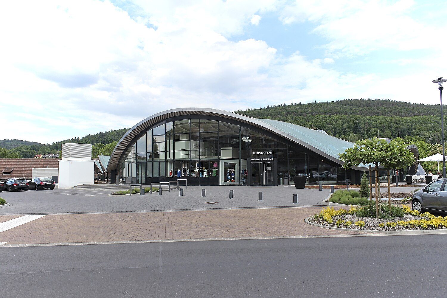 Die Toskana Therme Bad Orb von außen: geschwungenes Tonnendach mit Glasfront, davor ein Platz mit Blumenbeeten, im Hintergrund bewaldete Hügel