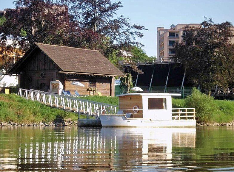 Saunaboot der Weser-Therme in Bad Karlshafen: weißes Boot am Steg auf der Weser, dahinter ein Holzhaus unter Bäumen